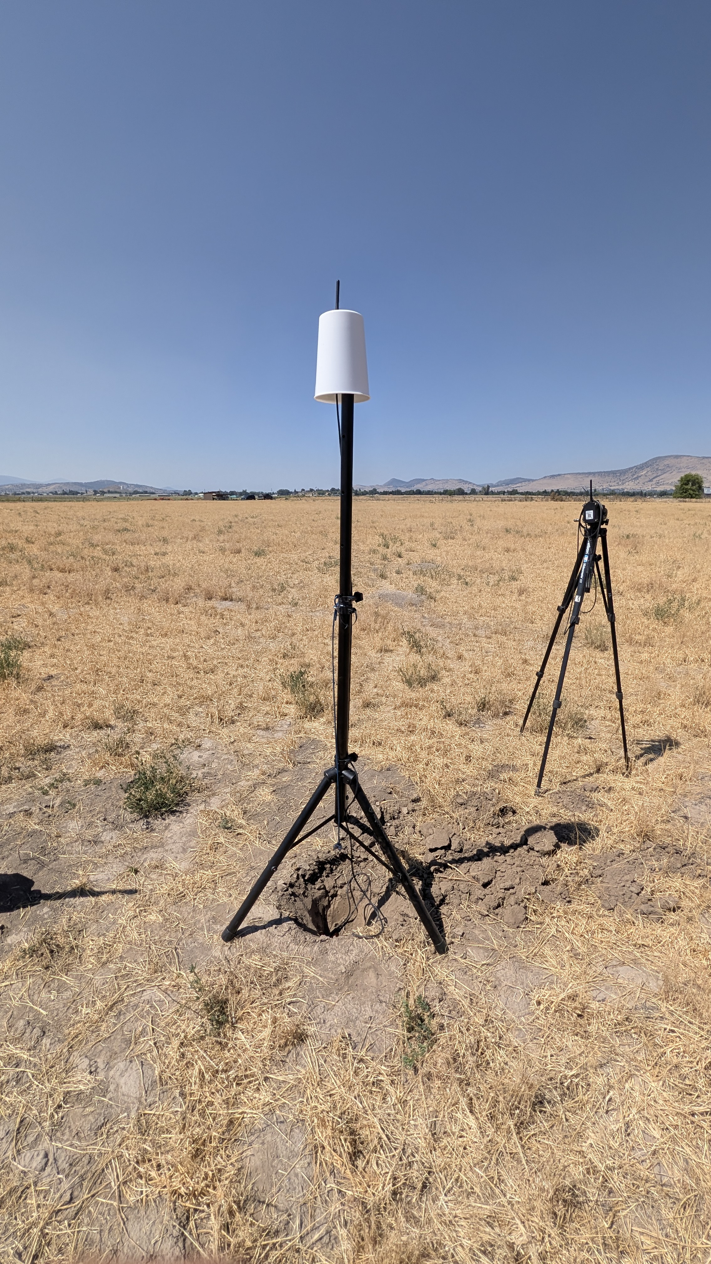 Basin Roots sensor and transmitter pole standing in an open field, with sky and the Klamath Basin mountains visible in the background